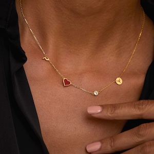 Pode incluir: Um colar de ouro com uma corrente delicada. O colar apresenta um pingente de coração vermelho, uma pequena gema transparente, um pingente circular com o símbolo de Áries e a letra "P". O colar é usado por uma pessoa.