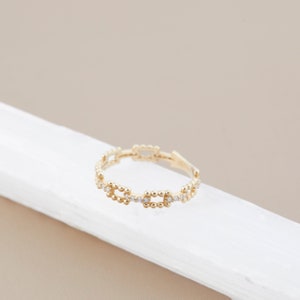 K&ouml;nnte beinhalten: Ein goldener Ring mit einem filigranen Kettenmuster, das mit kleinen, runden Steinen besetzt ist. Der Ring liegt auf einer wei&szlig;en Oberfl&auml;che.