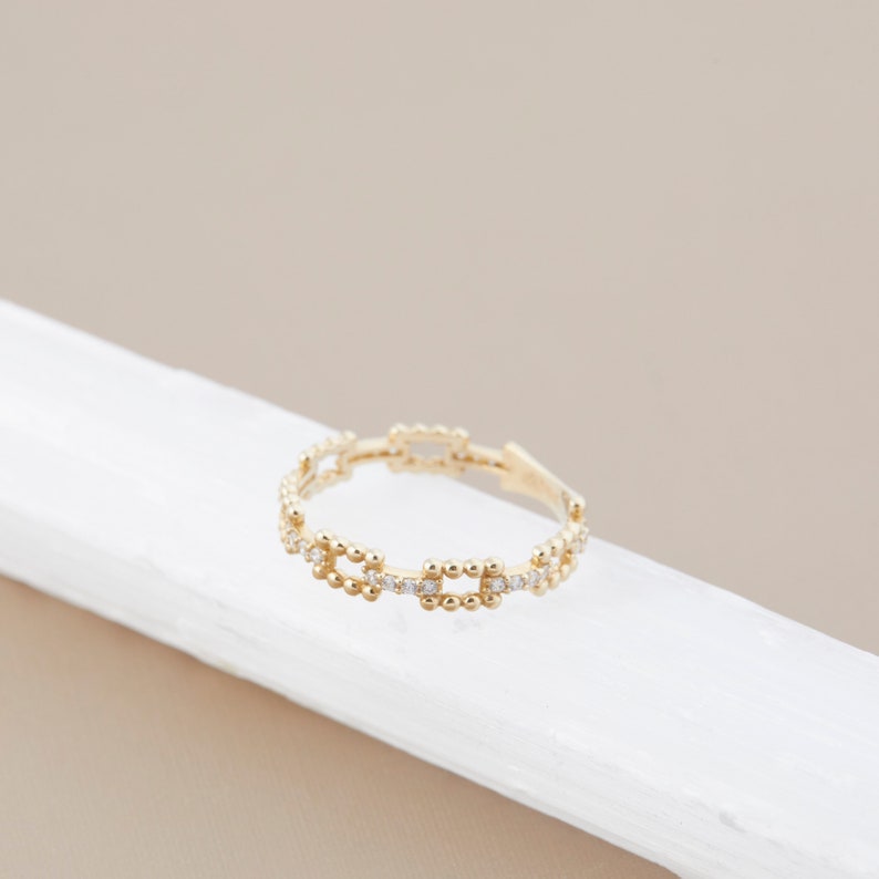 K&ouml;nnte beinhalten: Ein goldener Ring mit einem filigranen Kettenmuster, das mit kleinen, runden Steinen besetzt ist. Der Ring liegt auf einer wei&szlig;en Oberfl&auml;che.