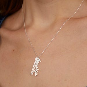 Könnte beinhalten: Eine silberfarbene vertikale Anhängerkette mit den Namen Jessica und Natalie in einer kursiven Schriftart. Der Anhänger hängt an einer zarten, dünnen silberfarbenen Kette, die auf der Haut aufliegt und einen schlichten Look erzeugt.