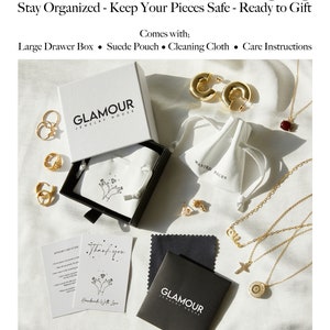 May include: A black box with the words "GLAMOUR JEWELRY HOUSE" on it. The box is open and contains a white pouch with the words "WANTED PIECES" on it. There are also several gold necklaces, earrings, and rings in the image. The image also includes a black cleaning cloth with the words "GLAMOUR JEWELRY HOUSE" on it and a card that says "Thank you. Handmade with love."