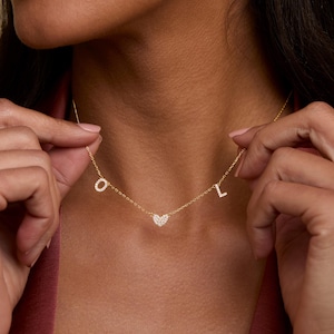 May include: A gold-toned chain necklace featuring three small, dangling charms: a letter O, a heart shape, and a letter L. Each charm is encrusted with small, clear, sparkling stones that catch the light, suspended from a delicate gold-colored link chain.