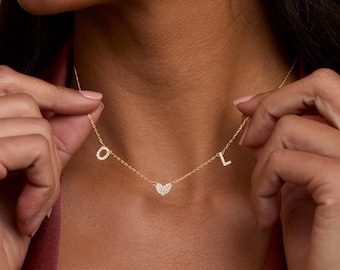 Puede incluir: Un collar de cadena en tono dorado con tres pequeños dijes colgantes: una letra O, una forma de corazón y una letra L. Cada dije está incrustado con pequeñas piedras claras y brillantes que captan la luz, suspendidos de una delicada cadena.