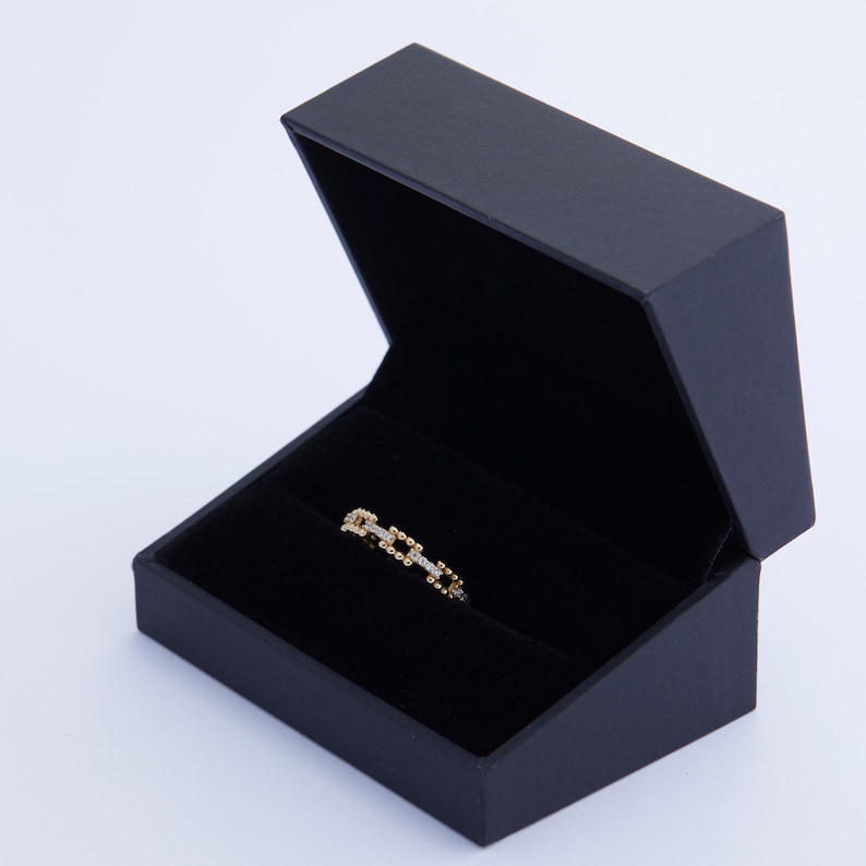K&ouml;nnte beinhalten: Ein goldener Ring mit einem quadratischen Kettenmuster und kleinen Diamanten, die in das Metall gefasst sind. Der Ring wird in einer schwarzen Samt-Ringbox pr&auml;sentiert.
