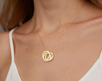 May include: A gold-toned necklace featuring three interlocking circular rings as a pendant. Each ring is engraved with text and hangs from a delicate gold-toned chain. The pendant rests against the collarbone, showcasing a polished metallic finish.