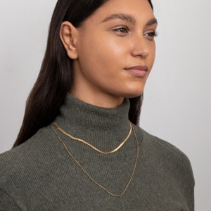 May include: A woman wearing a green turtleneck sweater with two gold chain necklaces. The necklaces are layered, with a thicker chain on top and a thinner chain underneath.