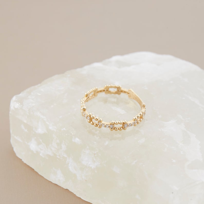 K&ouml;nnte beinhalten: Ein zarter Goldring mit einem Perlenmuster und kleinen klaren Steinen. Der Ring liegt auf einer wei&szlig;en Oberfl&auml;che.