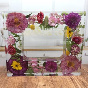 May include: A clear rectangular picture frame with a pressed flower design. The frame is filled with various dried flowers in shades of pink, purple, yellow, and red.