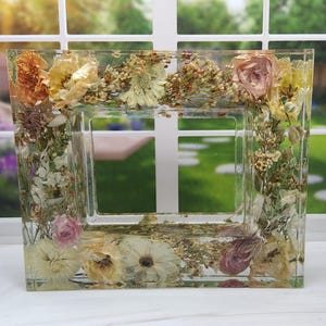 May include: A clear resin picture frame with pressed flowers and leaves in shades of pink, white, and yellow.