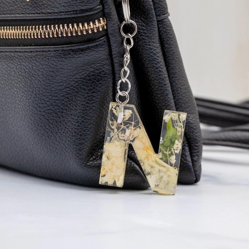 K&ouml;nnte beinhalten: Ein klarer Buchstabenanh&auml;nger "N" aus Kunstharz mit getrockneten Blumen. Der Schl&uuml;sselanh&auml;nger ist an einer silbernen Kette befestigt und h&auml;ngt vor einer schwarzen Ledertasche mit goldenem Rei&szlig;verschluss.