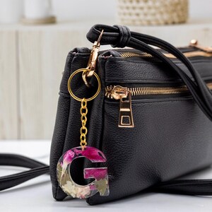 K&ouml;nnte beinhalten: Eine schwarze Umh&auml;ngetasche mit goldfarbener Hardware und einem floralen Buchstabenanh&auml;nger "G" aus Kunstharz. Der Anh&auml;nger zeigt rosa und wei&szlig;e Blumen, die in klarem Kunstharz eingeschlossen sind.
