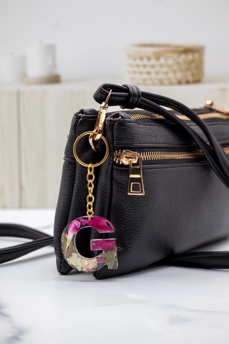 K&ouml;nnte beinhalten: Eine schwarze Umh&auml;ngetasche mit goldfarbener Hardware und einem floralen Buchstabenanh&auml;nger "G" aus Kunstharz. Der Anh&auml;nger zeigt rosa und wei&szlig;e Blumen, die in klarem Kunstharz eingeschlossen sind.