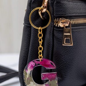K&ouml;nnte beinhalten: Ein Schl&uuml;sselanh&auml;nger mit dem Buchstaben "G" aus klarem Harz, gef&uuml;llt mit getrockneten rosa und wei&szlig;en Blumen. Die goldfarbene Kette und der Schl&uuml;sselring sind an einer schwarzen Ledertasche mit goldenem Rei&szlig;verschluss befestigt.