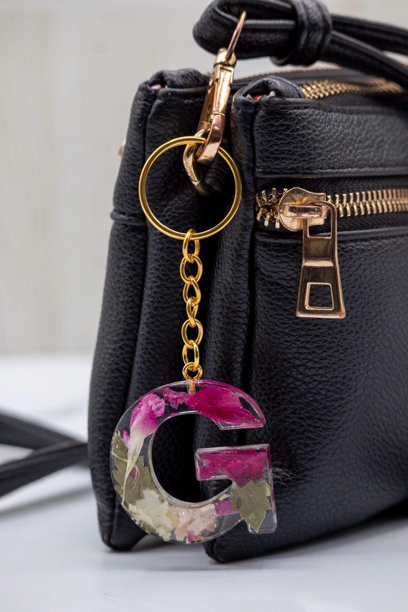 K&ouml;nnte beinhalten: Ein Schl&uuml;sselanh&auml;nger mit dem Buchstaben "G" aus klarem Harz, gef&uuml;llt mit getrockneten rosa und wei&szlig;en Blumen. Die goldfarbene Kette und der Schl&uuml;sselring sind an einer schwarzen Ledertasche mit goldenem Rei&szlig;verschluss befestigt.