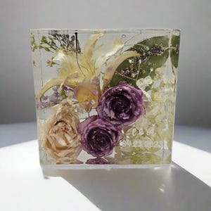 May include: A clear resin block encases dried flowers in shades of purple, cream, and yellow. The arrangement includes roses, lilies, and other delicate blooms. The block has a square shape and contains the engraved initials "C & A" and the date "04.23.2023".