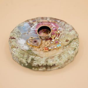 May include: A round, faceted resin tray embedded with dried white and pink flowers. Resting on the tray is a gold-tone heart-shaped pendant necklace with a dried flower inside, a gold-tone chain necklace with colorful floral charms, and a ring.