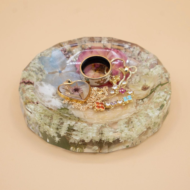 May include: A round, faceted resin tray embedded with dried white and pink flowers. Resting on the tray is a gold-tone heart-shaped pendant necklace with a dried flower inside, a gold-tone chain necklace with colorful floral charms, and a ring.