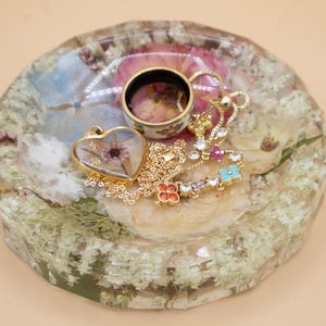 May include: A round, faceted resin tray containing embedded dried flowers in shades of white, pink, and blue. Resting on the tray is a gold-toned heart-shaped pendant with a dried flower inside, a gold chain, and a ring with a floral pattern.
