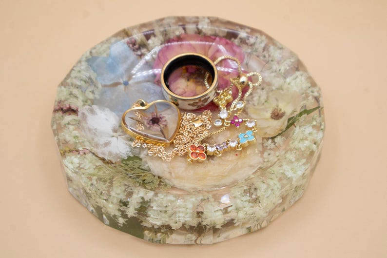 May include: A round, faceted resin tray containing embedded dried flowers in shades of white, pink, and blue. Resting on the tray is a gold-toned heart-shaped pendant with a dried flower inside, a gold chain, and a ring with a floral pattern.