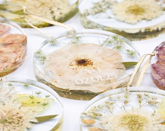 May include: Circular resin ornaments with dried flowers and gold ribbon. Some have initials and a date in white text. The flowers are in shades of cream, white, and purple. Each is a unique design.