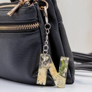 K&ouml;nnte beinhalten: Ein klarer Harz-Buchstabenanh&auml;nger "N" mit getrockneten Blumen und Goldflocken, der an einer silbernen Kette an einer schwarzen Ledertasche befestigt ist. Ein einzigartiges Accessoire.