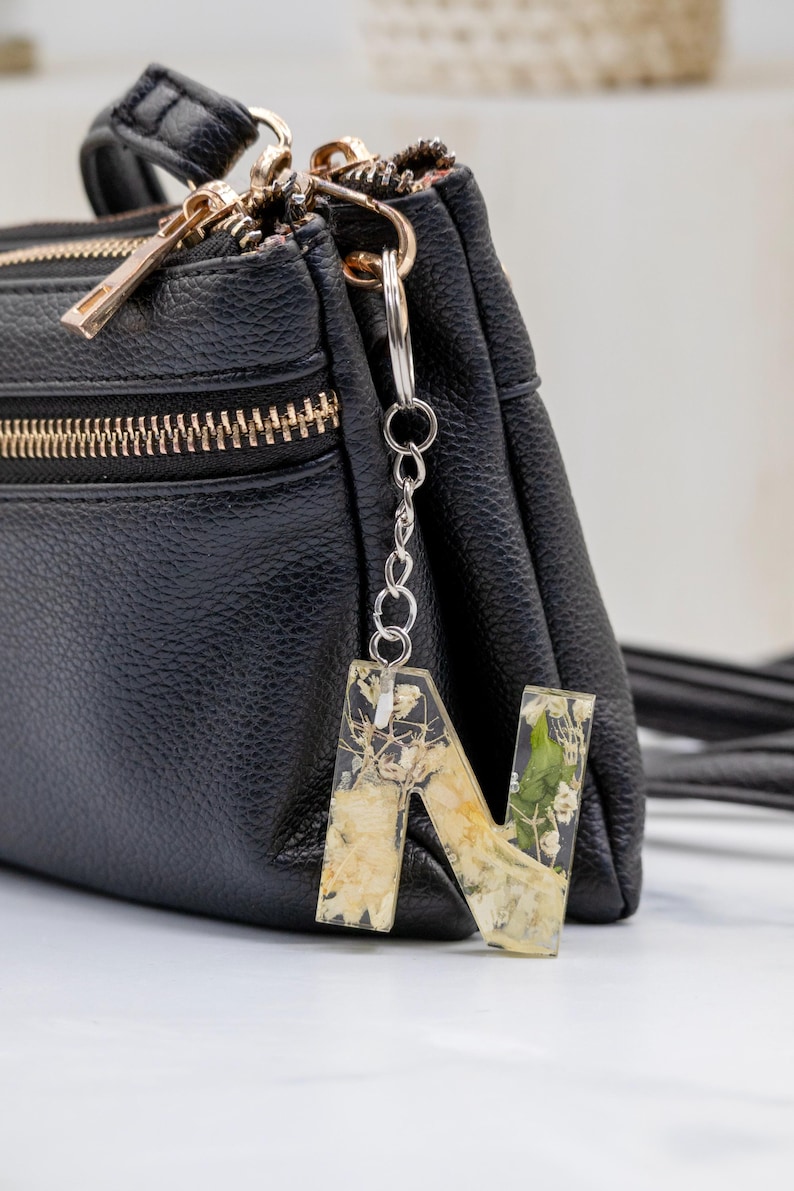 K&ouml;nnte beinhalten: Ein klarer Harz-Buchstabenanh&auml;nger "N" mit getrockneten Blumen und Goldflocken, der an einer silbernen Kette an einer schwarzen Ledertasche befestigt ist. Ein einzigartiges Accessoire.