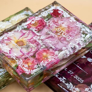 May include: Three rectangular clear resin blocks containing preserved flowers and white printed text. The center block features pink roses, a small crucifix, and the text Our Love Story, Suhail & Renee, November 28, 2025, with a white ornate border.