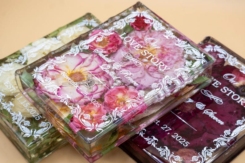 May include: Three rectangular clear resin blocks containing preserved flowers and white printed text. The center block features pink roses, a small crucifix, and the text Our Love Story, Suhail & Renee, November 28, 2025, with a white ornate border.