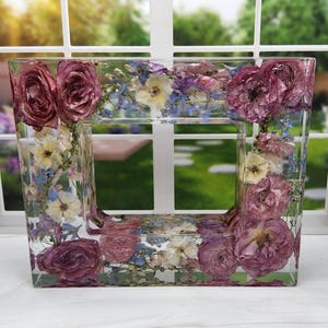 May include: A clear rectangular picture frame with a border of pressed flowers. The flowers are a mix of pink, white, and blue.