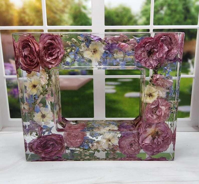 May include: A clear rectangular picture frame with a border of pressed flowers. The flowers are a mix of pink, white, and blue.