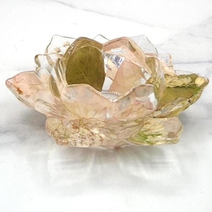May include: A clear, iridescent resin candle holder shaped like a flower. The petals are a soft pink with embedded dried leaves and flowers. The center is open to hold a candle, creating a unique decorative piece.