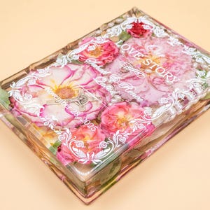 May include: A rectangular clear resin block containing preserved pink and red roses, white pearls, and a silver crucifix. The surface features white printed text reading Our Love Story, Sarah & Renee, and November 28, 2025, framed by a decorative border.