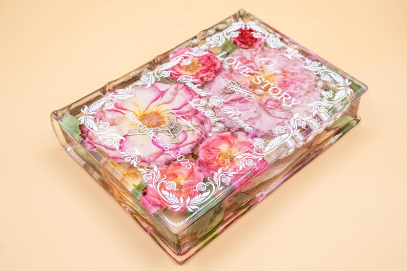 May include: A rectangular clear resin block containing preserved pink and red roses, white pearls, and a silver crucifix. The surface features white printed text reading Our Love Story, Sarah & Renee, and November 28, 2025, framed by a decorative border.