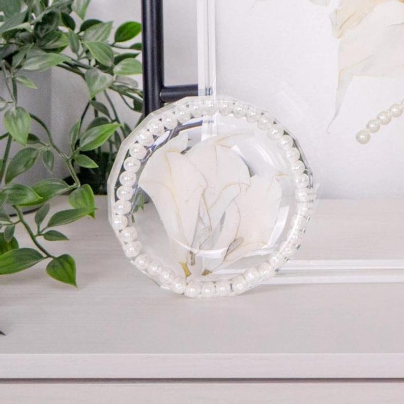 May include: A circular resin tray featuring a clear finish with embedded white flower petals. The outer edge of the tray is decorated with a border of small, round, white faux pearls arranged in a continuous ring around the perimeter.