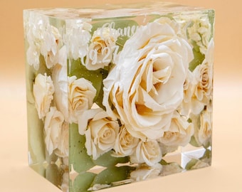 May include: A rectangular clear resin block containing preserved cream-colored roses and green eucalyptus leaves. The front surface features white engraved text that reads Chaney and 09.27. The floral arrangement is suspended within the solid material.