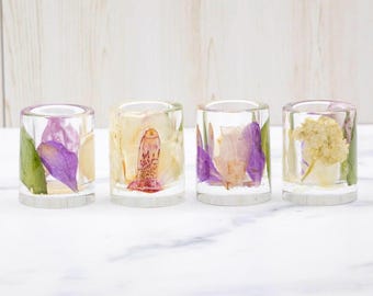 May include: Four clear resin votive candle holders, each containing a different pressed flower arrangement. The flowers include purple, yellow, and white petals and green leaves. The holders sit on a white marble surface.
