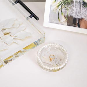 May include: A round, clear resin trinket dish featuring embedded white flower petals and a border of small white pearls. A thin gold ring with a small red stone charm rests in the center of the dish, which sits near a rectangular resin tray.