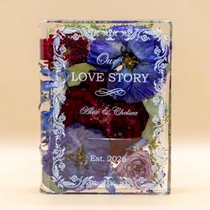 May include: A rectangular clear resin block containing pressed blue, purple, and deep red flowers. The front features white printed text reading Our LOVE STORY, Alex & Chelsea, and Est. 2026, framed by an ornate white decorative border.