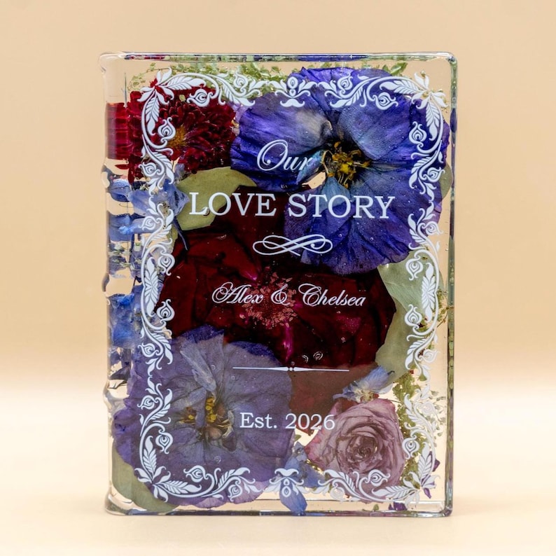 May include: A rectangular clear resin block containing pressed blue, purple, and deep red flowers. The front features white printed text reading Our LOVE STORY, Alex & Chelsea, and Est. 2026, framed by an ornate white decorative border.