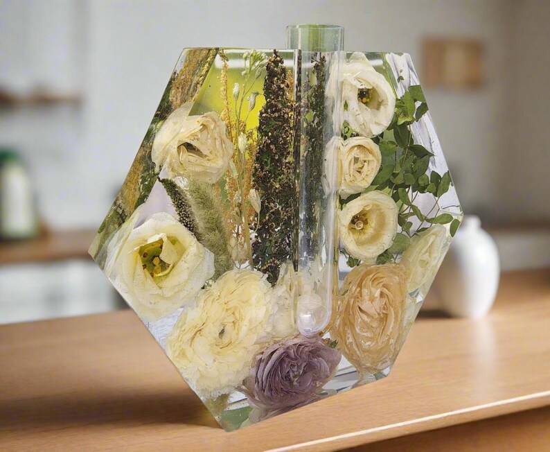 K&ouml;nnte beinhalten: Eine sechseckige, transparente Harzvase mit getrockneten Blumen und Gr&uuml;npflanzen im Inneren. Die Vase hat ein transparentes Rohr in der Mitte, um Wasser zu halten.