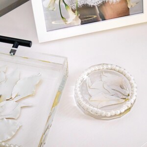 May include: A round, clear resin coaster or trinket dish featuring embedded white flower petals and a border of small, white, spherical faux pearls. The piece is positioned next to a rectangular clear tray with similar floral accents.
