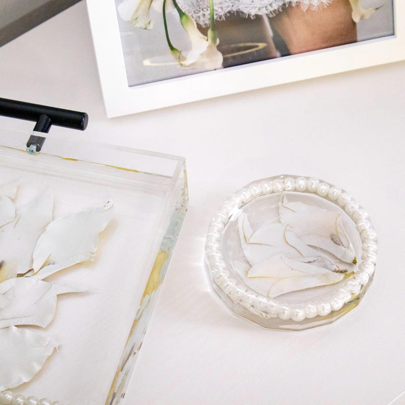 May include: A round, clear resin coaster or trinket dish featuring embedded white flower petals and a border of small, white, spherical faux pearls. The piece is positioned next to a rectangular clear tray with similar floral accents.