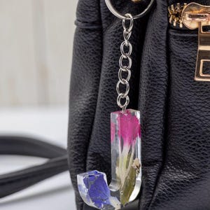 K&ouml;nnte beinhalten: Ein klarer Kunstharz-Schl&uuml;sselanh&auml;nger in Form des Buchstabens &bdquo;J&ldquo; mit eingebetteten rosa und blauen Blumen. Der Schl&uuml;sselanh&auml;nger ist an einer silbernen Kette und einem Ring befestigt, vor einer schwarzen Ledertasche.