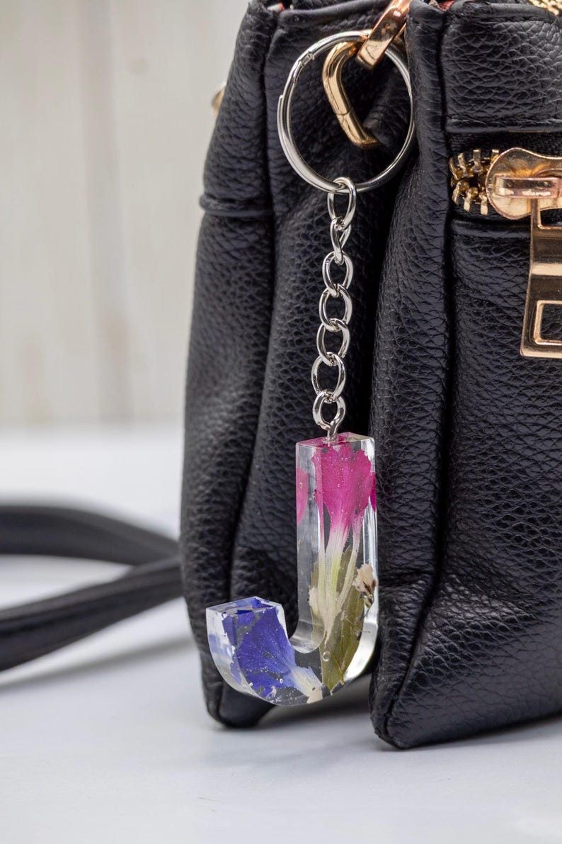 K&ouml;nnte beinhalten: Ein klarer Kunstharz-Schl&uuml;sselanh&auml;nger in Form des Buchstabens &bdquo;J&ldquo; mit eingebetteten rosa und blauen Blumen. Der Schl&uuml;sselanh&auml;nger ist an einer silbernen Kette und einem Ring befestigt, vor einer schwarzen Ledertasche.