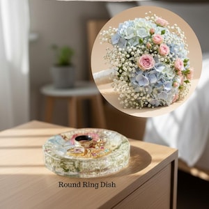 May include: A round resin ring dish containing embedded dried flowers and small jewelry pieces. Above, a circular inset shows a bridal bouquet of light blue hydrangeas, pink roses, and baby's breath. Text reads: Bouquet Transformation with resin flower preservation, Round Ring Dish, Save for Later, www.wildcoastflowerpreservation.com.