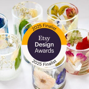 May include: A collection of clear cylindrical resin candle holders embedded with various pressed flowers and greenery. A circular badge in the center displays the text 2025 Finalist Etsy Design Awards 2025 Finalist in orange, white, and dark blue.
