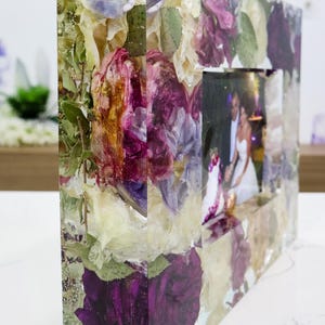May include: A clear resin picture frame with dried flowers and leaves embedded in the resin. The frame is rectangular and has a square opening for a photo. The flowers are in shades of purple, white, and green.