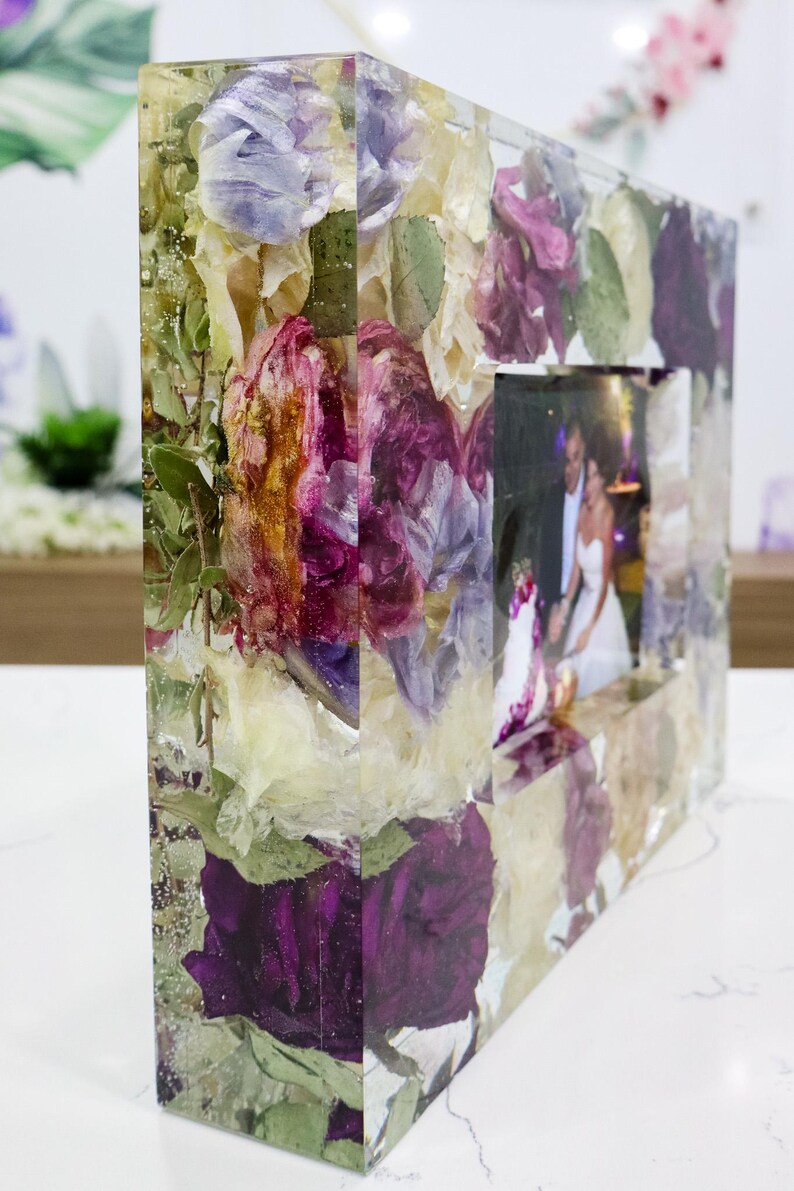 May include: A clear resin picture frame with dried flowers and leaves embedded in the resin. The frame is rectangular and has a square opening for a photo. The flowers are in shades of purple, white, and green.