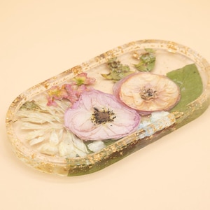 May include: An oval-shaped resin tray featuring embedded dried flowers in shades of pink, white, and green. The clear resin is accented with gold flakes throughout the rim and base, creating a decorative piece with a smooth, glossy finish.