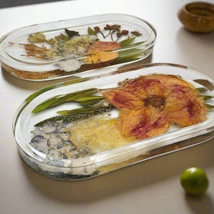 May include: Two oval-shaped clear resin trays featuring embedded dried flowers and greenery. The foreground tray displays a large pink and yellow flower, green leaves, and blue petals. The background tray contains various dried botanical elements.